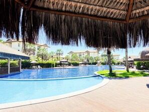 Ein Swimmingpool mit einer überdachten, mit Reet gedeckten Struktur, umgeben von Palmen und Liegestühlen.