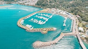 Vue aérienne d’un port de plaisance situé sur la côte, avec des bateaux amarrés, entouré d’eau et de verdure.