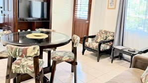 Un salon avec une table ronde en verre, des fauteuils à motifs floraux, un téléviseur posé sur une étagère en bois, une porte avec des stores et une fenêtre avec des rideaux.