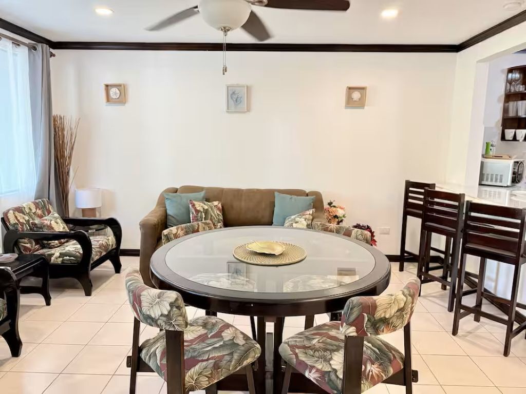 A dining area with a round glass table, floral-patterned chairs, a brown sofa, and a ceiling fan.