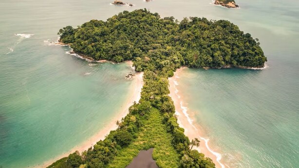 Vue aérienne d’une île tropicale, avec une étroite bande de plage séparant deux portions de terre plus importantes, le tout étant entouré d’eaux turquoise et cristallines.