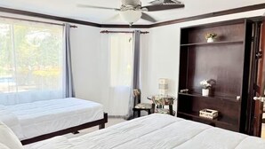 Une chambre d’hôtel avec deux lits, un ventilateur de plafond, une fenêtre avec des rideaux, une petite table avec une lampe et un buffet en bois foncé.