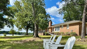 A row of white Adirondack chairs in a grassy area with a brick building and trees in the background. - Lake Murray home with boat ramp, dock, panoramic views close to USC@Columbia. (Chapin)