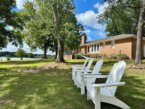 A row of white Adirondack chairs in a grassy area with a brick building and trees in the background. - Lake Murray home with boat ramp, dock, panoramic views close to USC@Columbia. (Chapin)