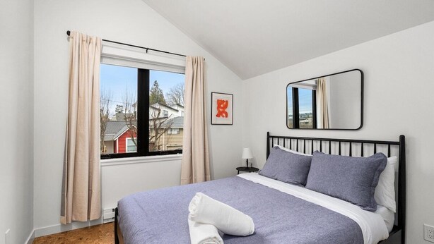 Un dormitorio con cama, ventana con cortinas, mesita de noche y espejo montado en la pared.