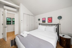 Une chambre à coucher comprenant un lit, une table de chevet, une lampe fixée au mur et une porte donnant sur une autre pièce.