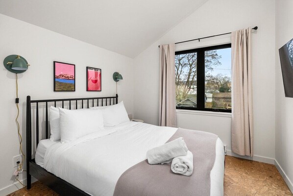 Un lit bien fait, avec du linge de lit blanc et une serviette enroulée, une fenêtre avec des rideaux et deux lampes fixées au mur.
