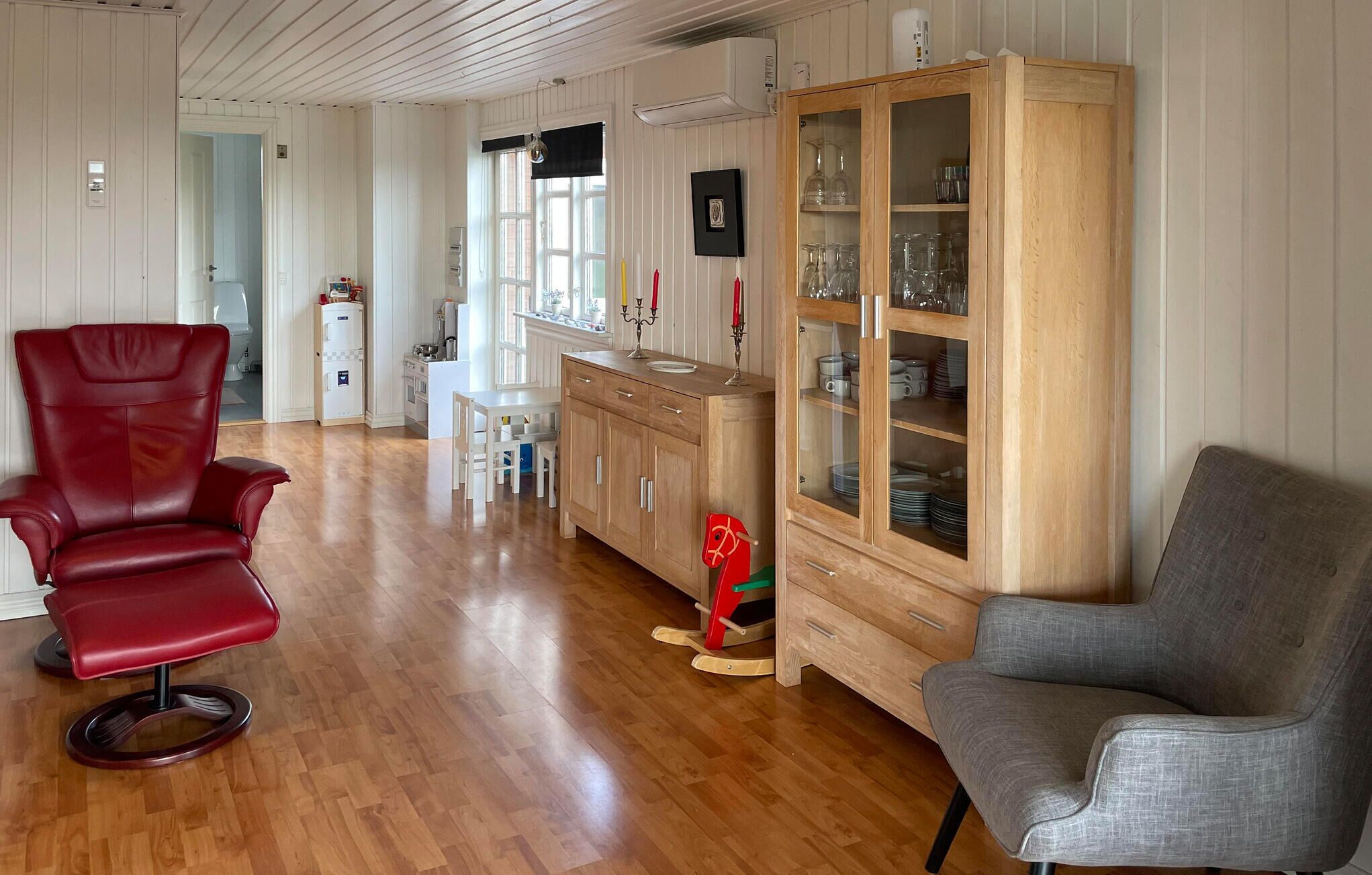 Une pièce avec un plancher en bois, une chaise rouge, une chaise grise, un meuble en bois doté de portes en verre et une petite table.