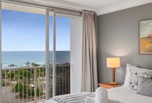 A hotel room with a bed, a nightstand with a lamp, a painting on the wall, and a view of the ocean through a sliding door. - Panoramic Beachfront Escape | 180° Ocean Views | Heated Pool & Spa (Sunshine Coast)