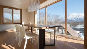 A modern dining area with a wooden table and chairs, large windows with a snowy mountain view, and a hanging light fixture.