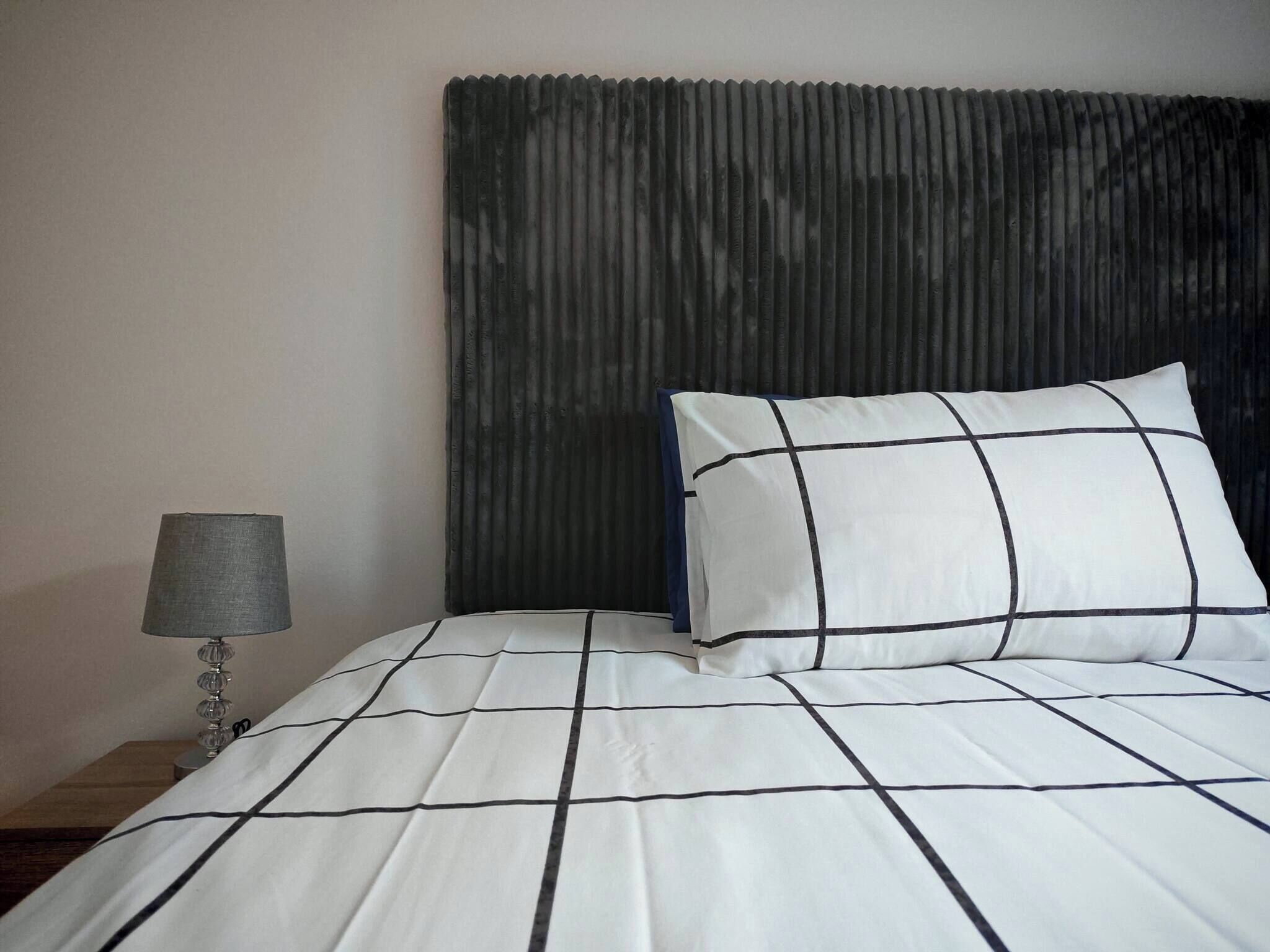 Ein Bett mit einer Bettdecke in Weiß und Schwarz mit Rautenmuster, ein Kissen in ähnlichem Muster, ein dunkles Kopfteil und eine Nachttischlampe mit grauem Lampenschirm.