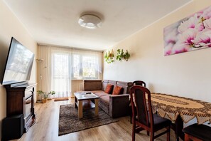 Ein Wohnzimmer mit einer Couch, einem Fernseher, einem Tisch und einem Fenster.