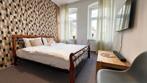 Un dormitorio con pared de estampado geométrico, una cama con sábanas blancas, una mesita de noche de madera, una silla y un televisor montado en la pared.