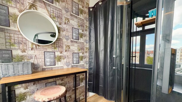 Una habitación con pared de ladrillo, un espejo redondo, una mesa de madera con un taburete y una cortina negra.