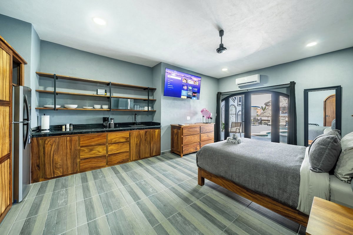 Habitación de hotel moderna con cama, kitchenette, televisor de pantalla plana y vista al área de la piscina.