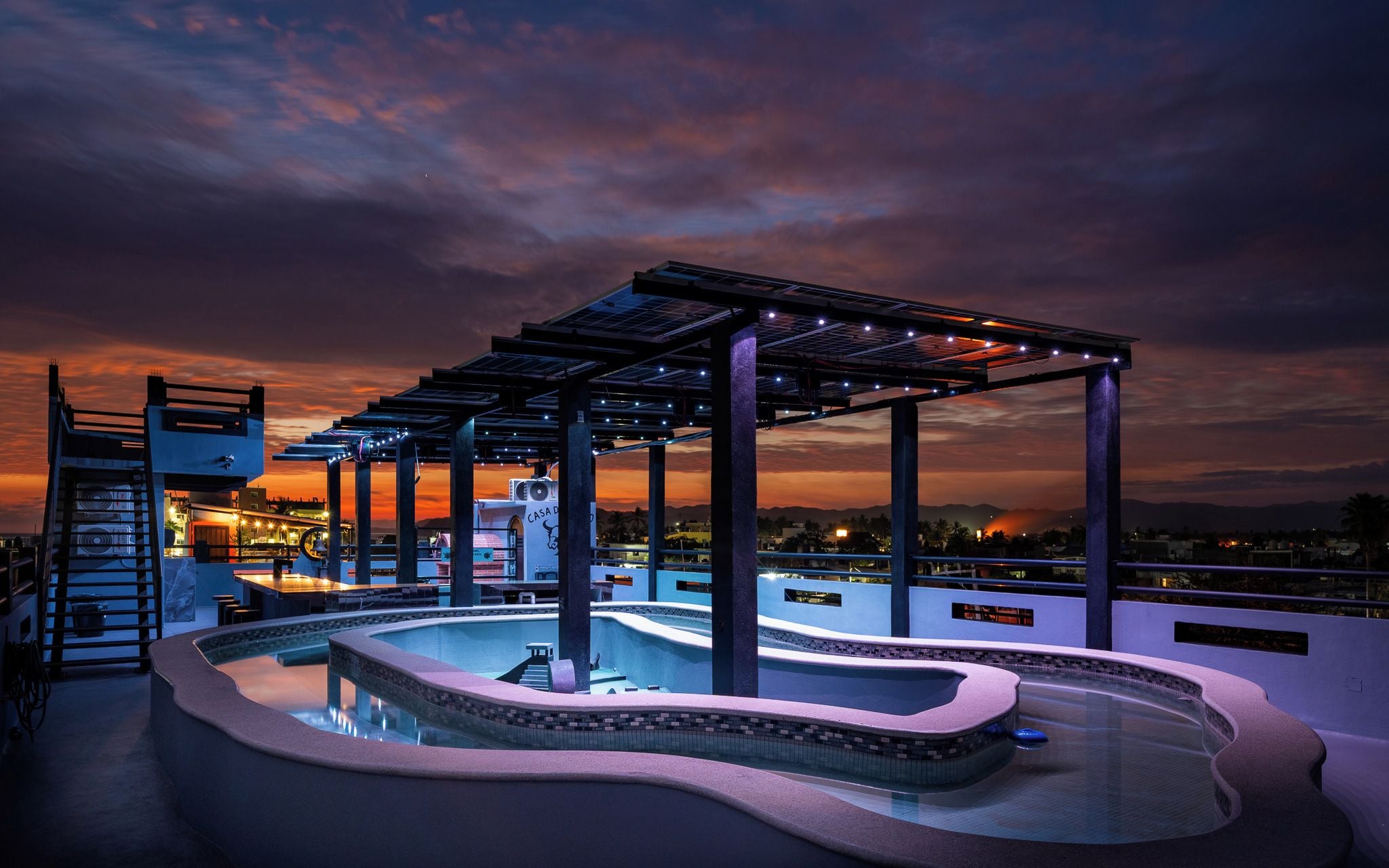A rooftop pool with a pergola, a hot tub, and a view of the cityscape at dusk.