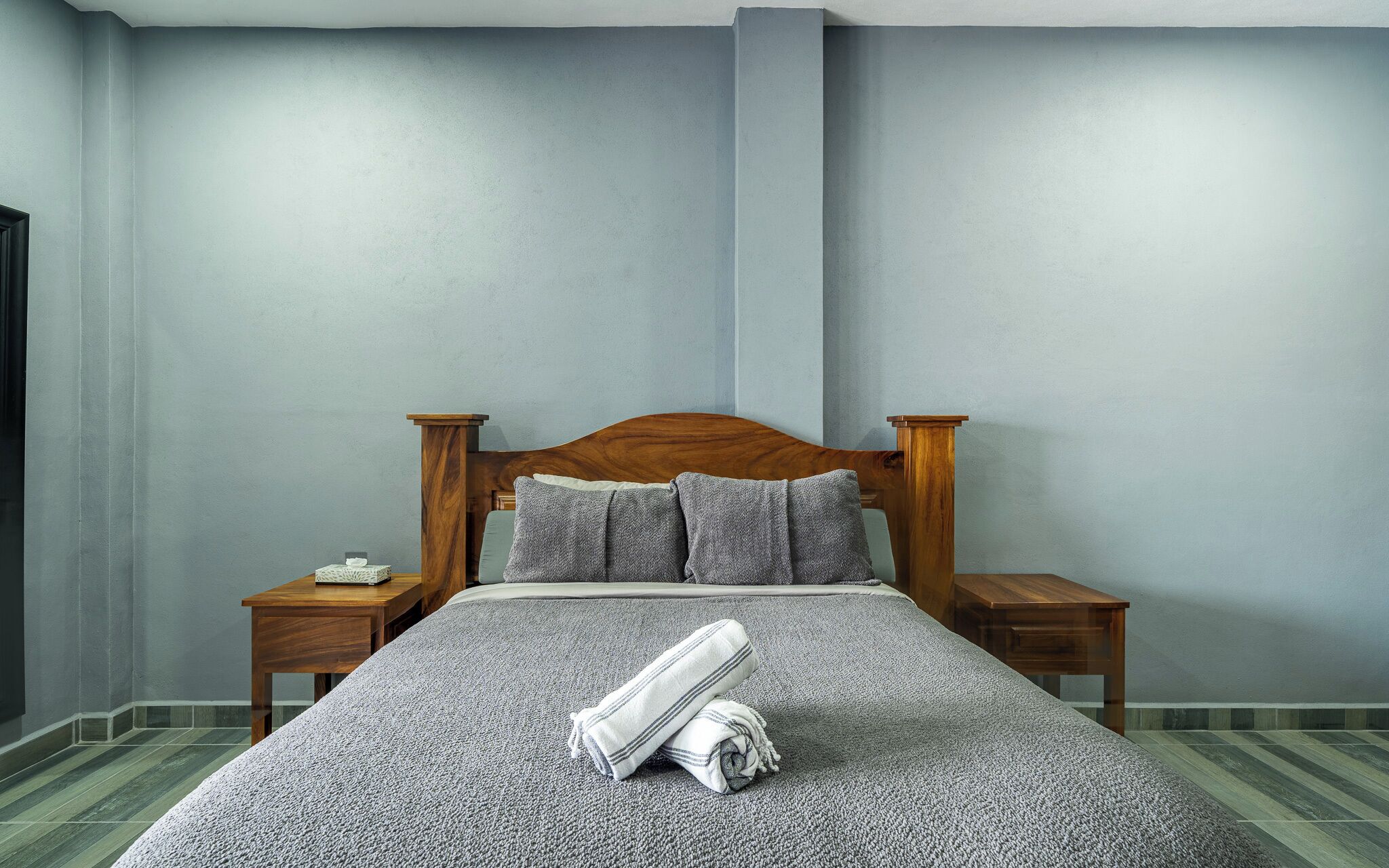 Un lit bien fait, recouvert d’une couverture grise à texture intéressante et agrémenté d’oreillers, avec deux tables de chevet en bois de chaque côté, dont l’une porte une petite lampe.