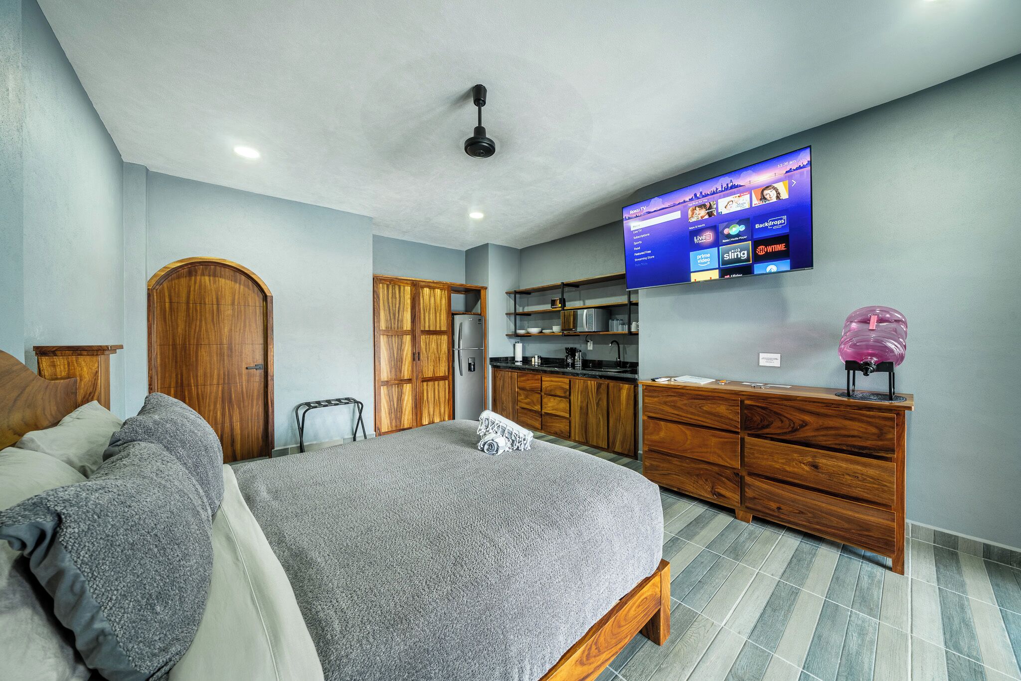 A bedroom with a bed, wooden wardrobe, a television mounted on the wall, and a kitchen area with cabinets and a refrigerator.
