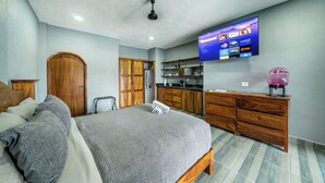 Un dormitorio con cama, armario de madera, televisor montado en la pared y una zona de cocina con armarios y refrigerador.