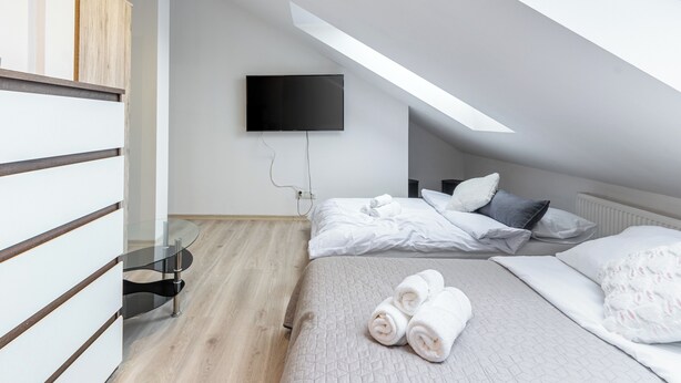 Ein modernes Schlafzimmer mit schrägverlaufender Decke, einem an der Wand montierten Flachbildfernseher, einem Bett mit weißer Bettwäsche und einem Glastisch.