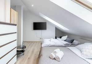 A modern bedroom with a sloped ceiling, a flat-screen TV mounted on the wall, a bed with white bedding, and a glass table. - Sikorskiego Comfort Studio #5 (Rzeszów)