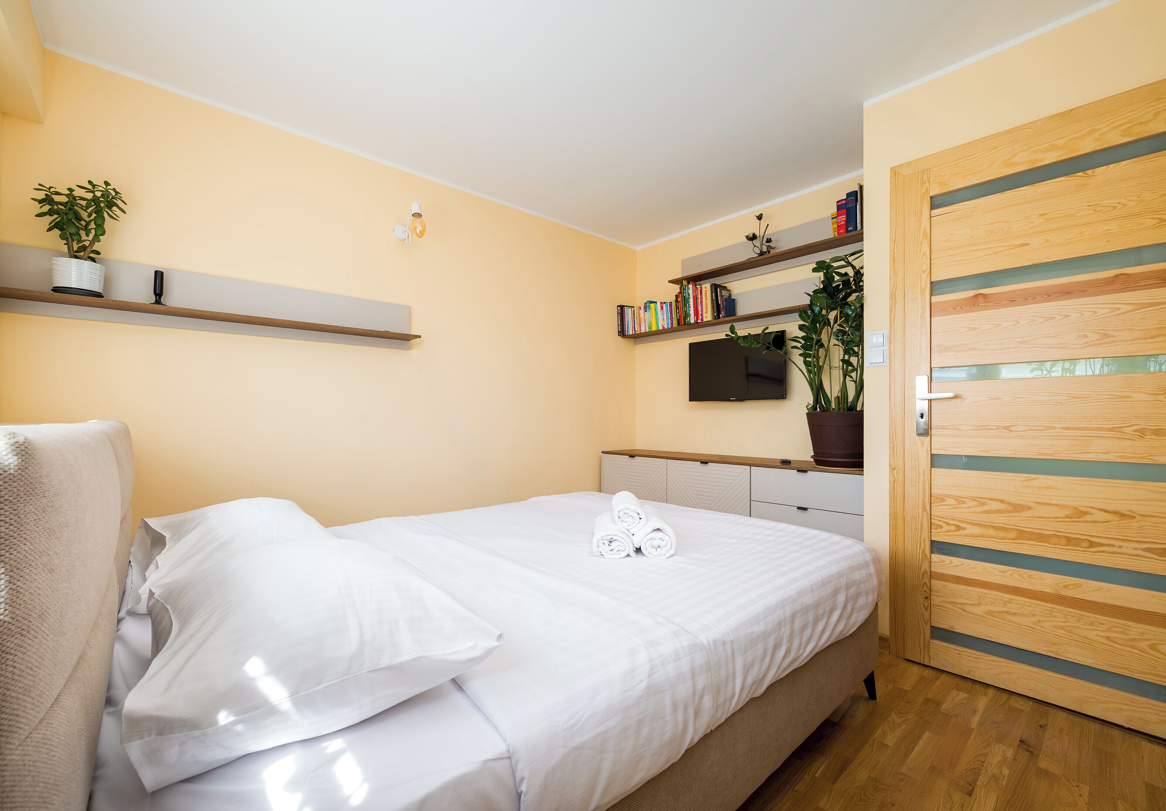 Un lit bien fait, avec du linge de lit blanc, une table de chevet, un téléviseur fixé au mur et une porte en bois.