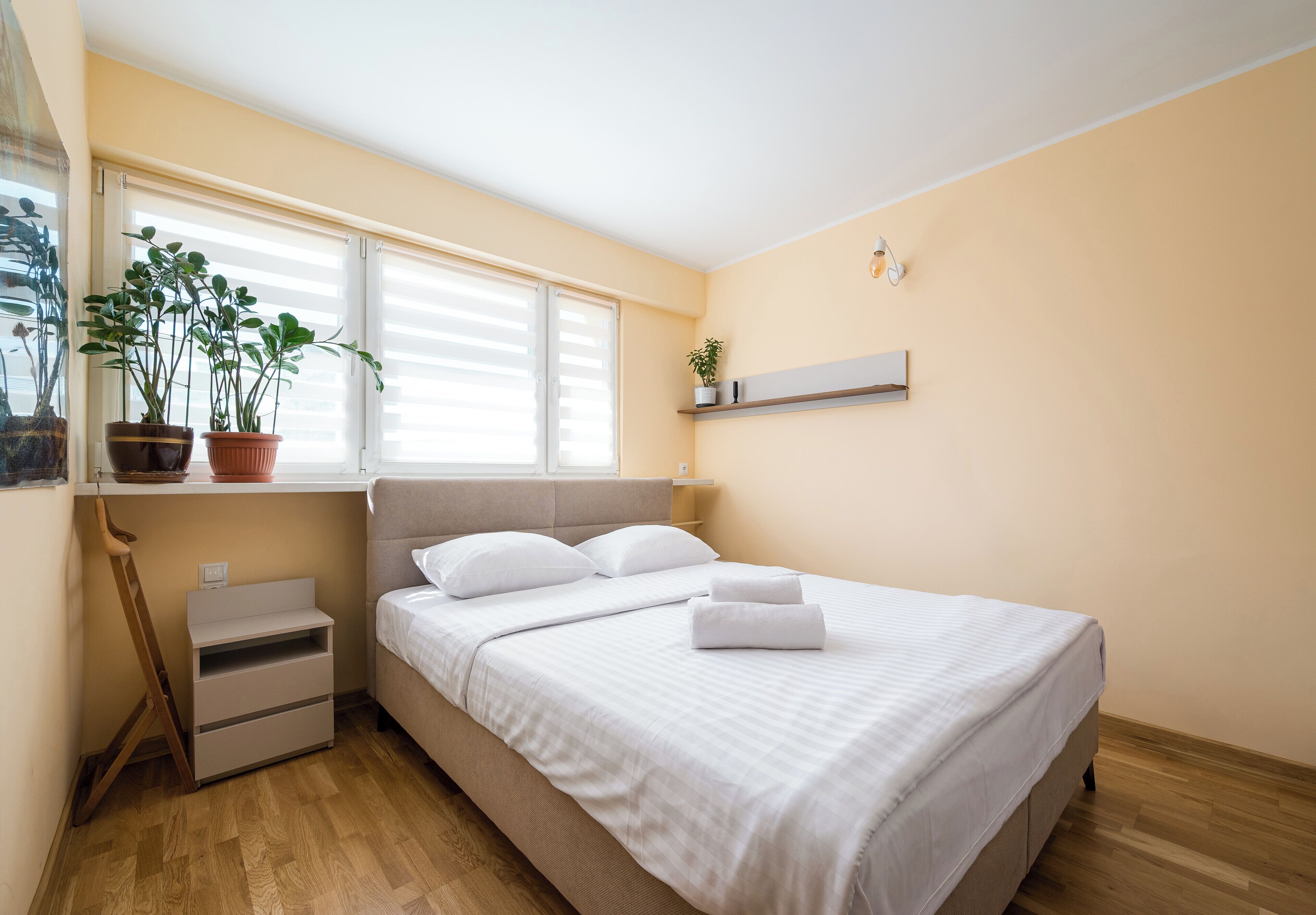 A bedroom with a bed, bedside tables, a window with blinds, a shelf with a plant, and a potted plant on the windowsill.