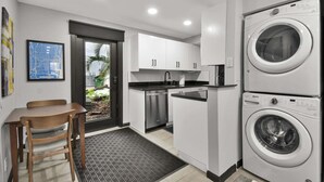 Une cuisine compacte équipée d’un lave-linge, un coin repas avec une chaise, et une porte donnant sur un balcon agrémenté de plantes.