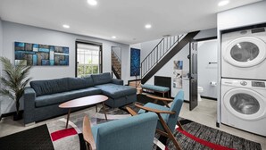 Een moderne woonkamer met een modulaire bank, een kleine ronde tafel en een blauwe fauteuil. Een trap leidt naar een hoger gelegen niveau, en er is een wasmachine zichtbaar op de achtergrond.