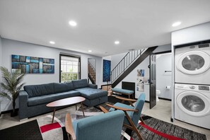 A modern living room with a sectional sofa, a small round table, and a blue armchair. There is a staircase leading to another level, and a washing machine is visible in the background. - Bright Designer Villa near Fort Lauderdale / Beach (Wilton Manors)