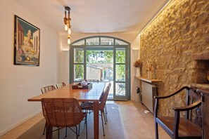 Un coin repas avec une longue table, des chaises en osier, un mur en pierre et une vue à travers une porte vitrée.