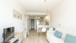 Un salon moderne avec un téléviseur à écran plat, un canapé blanc agrémenté de coussins turquoise, un coin repas avec une table et des chaises blanches, et une cuisine en arrière-plan.