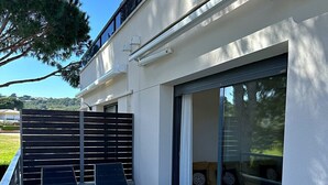 An outdoor patio with a glass table and white chairs, a sliding glass door, and a view of a forested area. - Sol, terraza y piscina en S’Agaró (Sagaro)