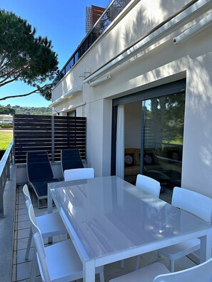Eine Außenpatio mit Glastisch und weißen Stühlen, eine Schiebetür und Blick auf einen bewaldeten Bereich.