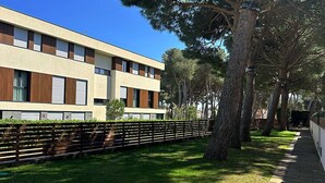 A modern building with a flat roof, large windows, and a paved pathway alongside a grassy area with trees. - Sol, terraza y piscina en S’Agaró (Sagaro)