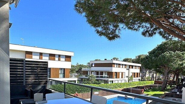 A balcony with a table and chairs, a pool, and modern buildings in the background. - Sol, terraza y piscina en S’Agaró (Sagaro)