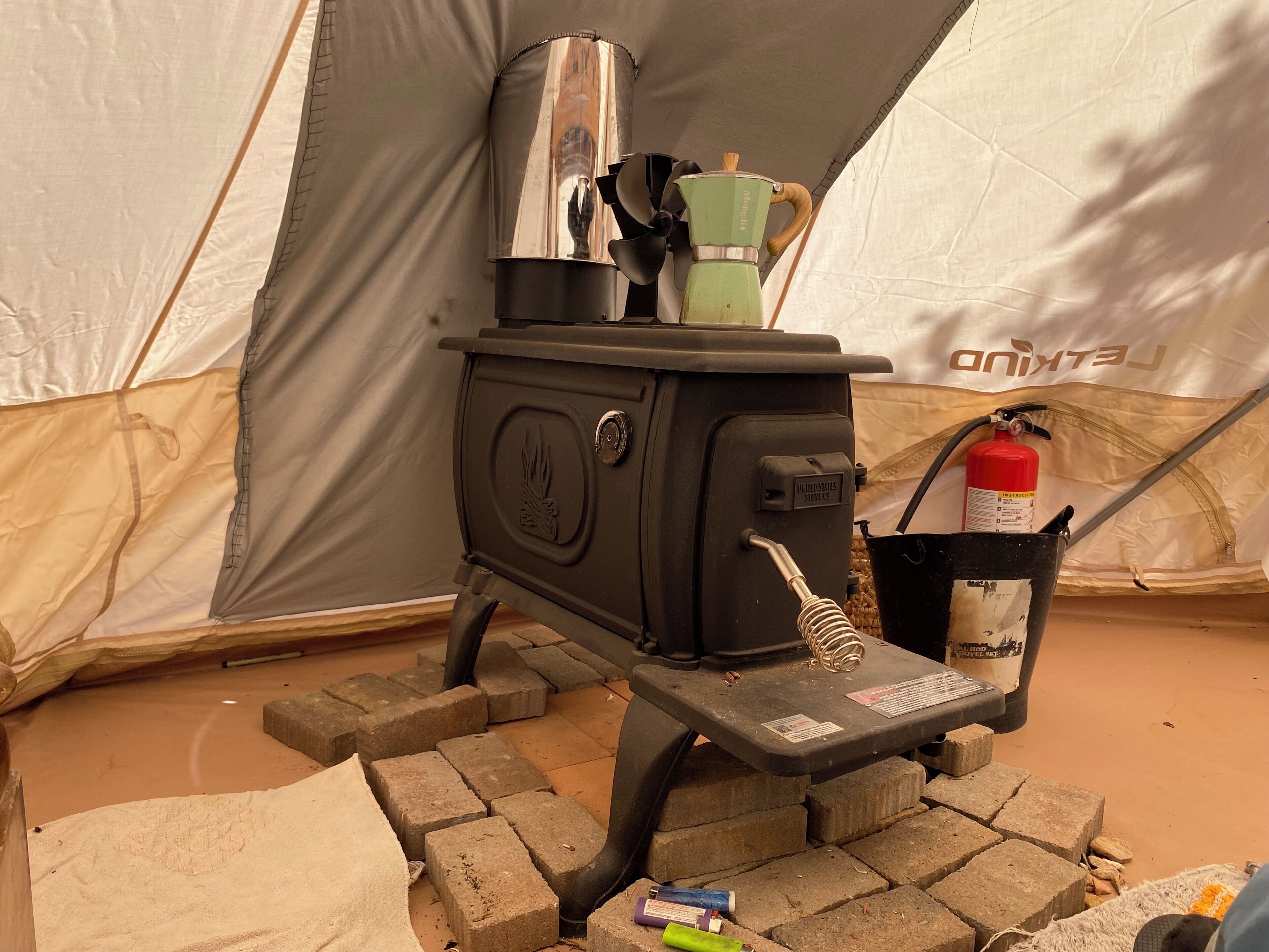 A portable stove with a kettle, a fire extinguisher, and a water bottle on a brick base.