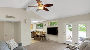 Una sala de estar con ventilador de techo, un sofá gris, una zona de comedor con mesa y sillas, y un televisor montado en la pared.