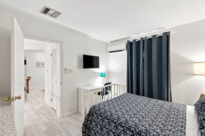 A bedroom with a bed, a desk with a lamp, a television, and a window with curtains.
