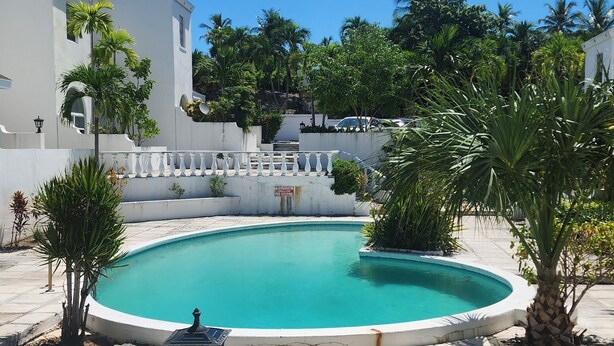Une piscine avec un mur blanc et un panneau, entourée de palmiers et d’un bâtiment blanc.