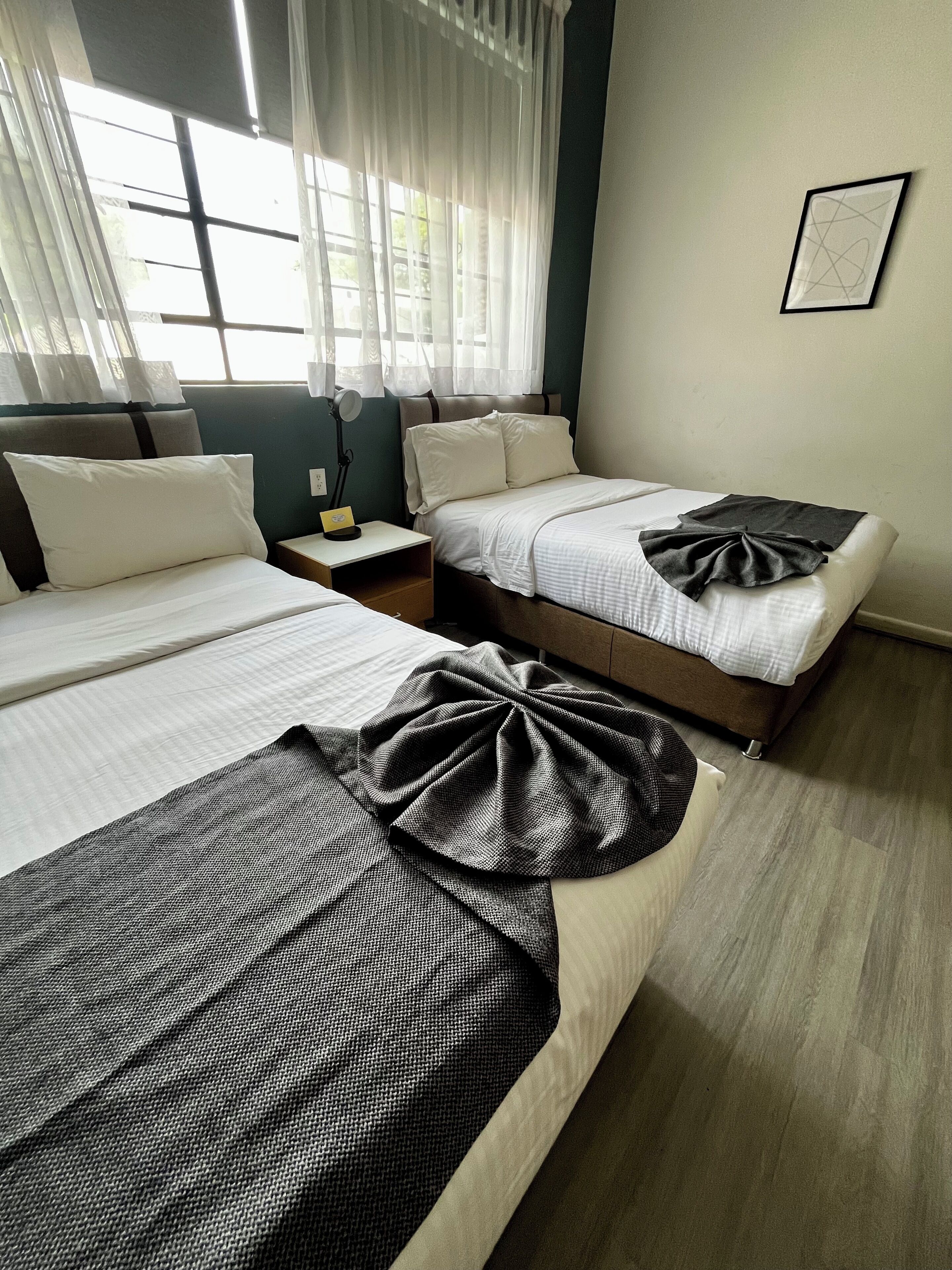 A hotel room with two beds, a bedside table with a lamp, a framed picture on the wall, and a window with sheer curtains.