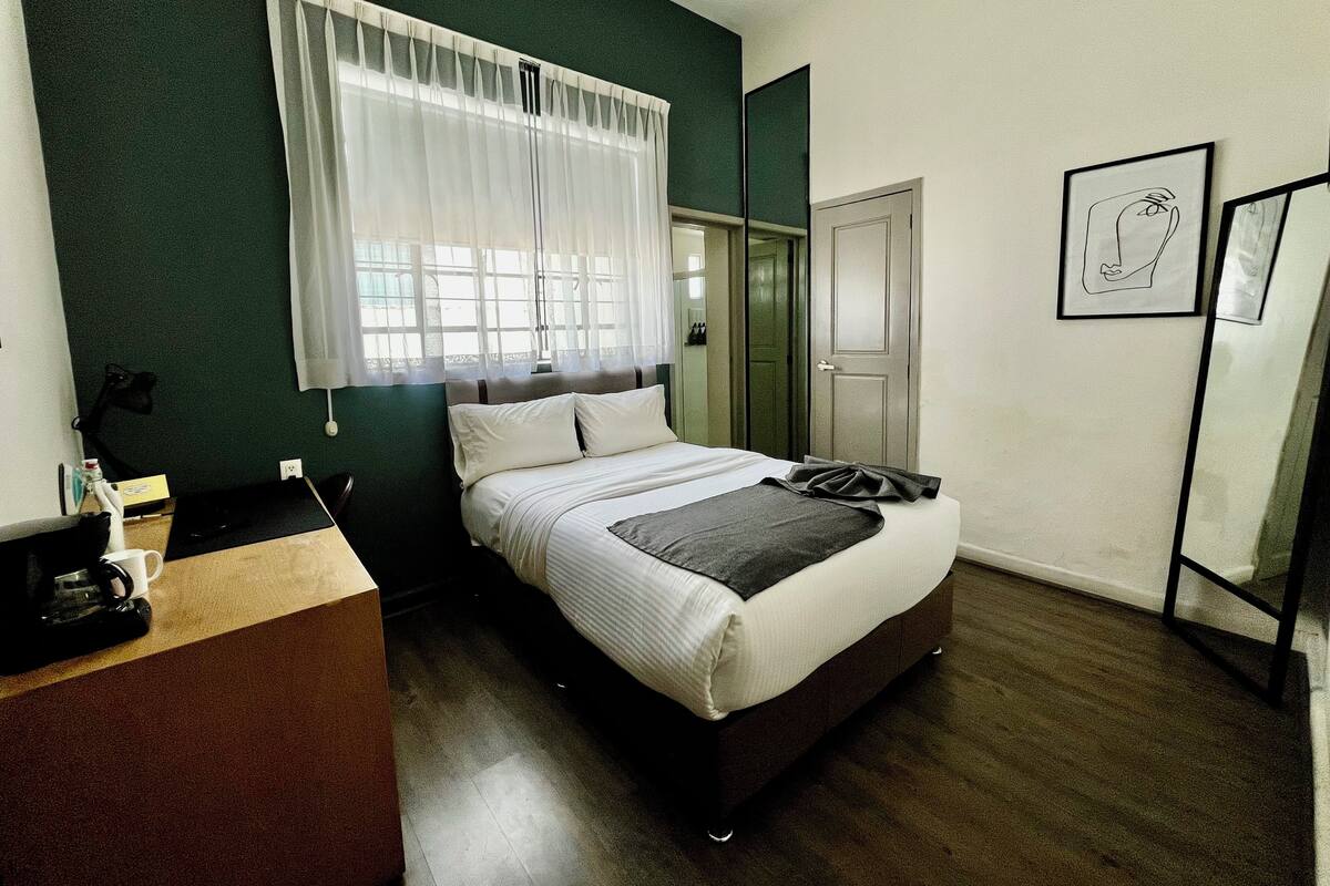 Habitació d'hotel amb un llit, una taula de treball, una cadira, un mirall i una porta.