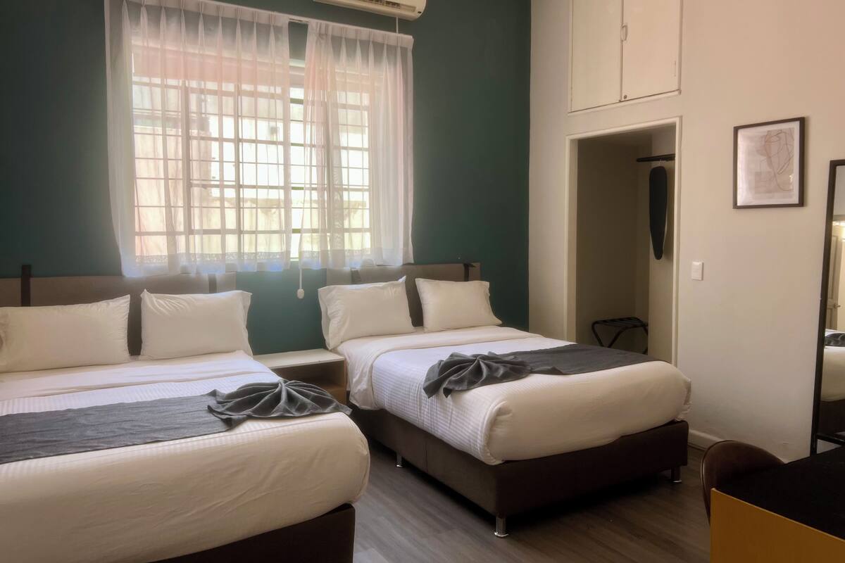 Habitació d'hotel amb dos llits, una finestra amb cortines lleugeres, un aparell d'aire condicionat, un armari i un quadre al mur.
