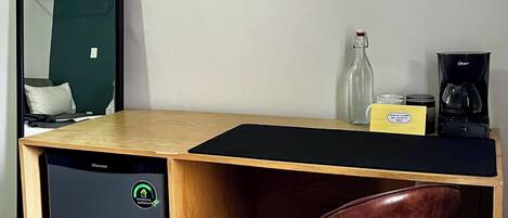 A compact, modern room with a desk, a compact refrigerator, a chair, a mirror, and a coffee maker.
