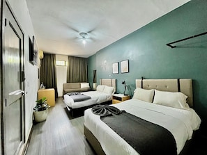 Habitación de hotel con dos camas, un ventilador de techo, un ventanal grande con cortinas, una planta en maceta y un televisor de pantalla plana.