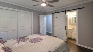 Een slaapkamer met een bed, een plafondventilator en een badkamer met een wastafel en spiegel.
