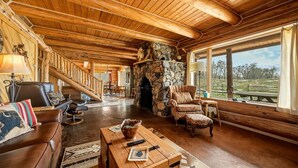 A log cabin interior with a stone fireplace, wooden ceiling beams, a staircase, and a large window offering a view of the outdoors.