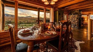 A wooden dining room with a table set for four, a stone fireplace, and a view of a mountainous landscape.