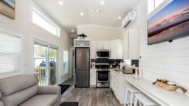 A modern kitchen with white cabinetry, a stainless steel refrigerator, a microwave, an oven, and a sink. There is a dining area with a white table and bar stools. - Near Beach • Pool + Amenities • Sleeps 8 (Middle Township)