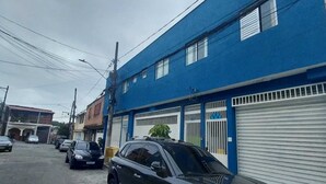A street with a blue building, parked cars, and a closed garage door. - Cozy apartment in Interlagos (São Paulo)